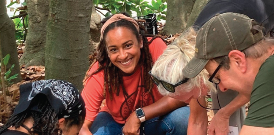 A group of adults gathered around the earth, examining the ground with interest, as one woman smiles directly at the camera, capturing a moment of shared discovery and joy.