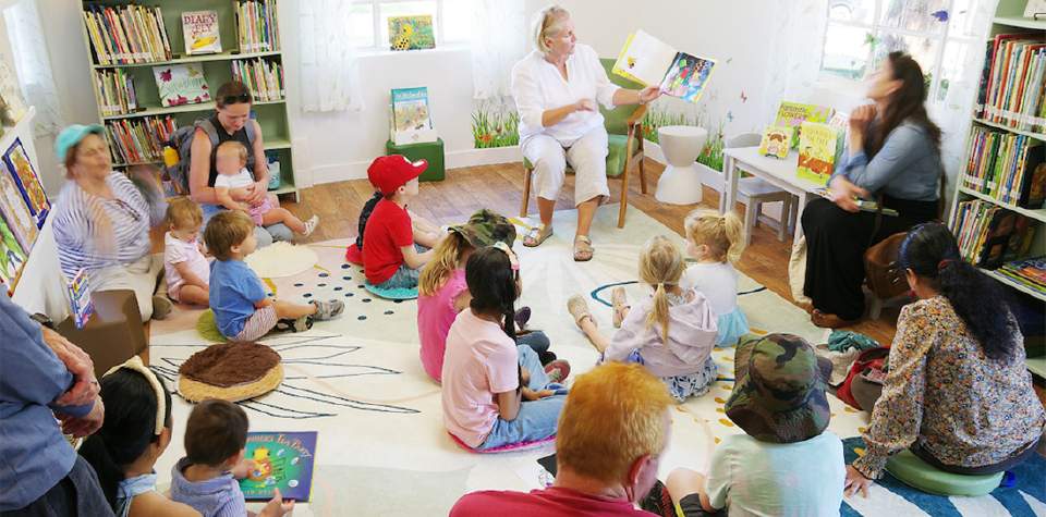 Young children gathered on the carpet of the brightly lit Children's Nature Library at River Farm, captivated by a woman reading a book during Story Time, surrounded by nature-themed books and educational resources.