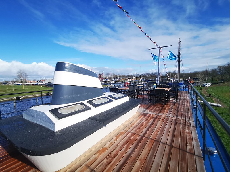 The patio on the Magnifique IV, a luxury river barge, features a partially-covered teak sundeck with tables and chairs. This inviting outdoor space allows passengers to enjoy the scenic views of the Dutch waterways while relaxing in comfort.