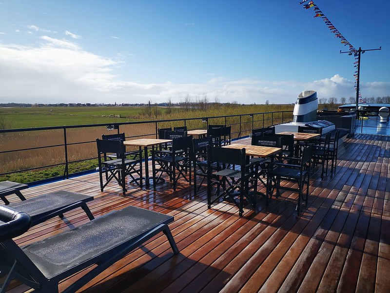 The patio on the Magnifique IV, a luxury river barge, features a partially-covered teak sundeck with tables and chairs. This inviting outdoor space allows passengers to enjoy the scenic views of the Dutch waterways while relaxing in comfort.