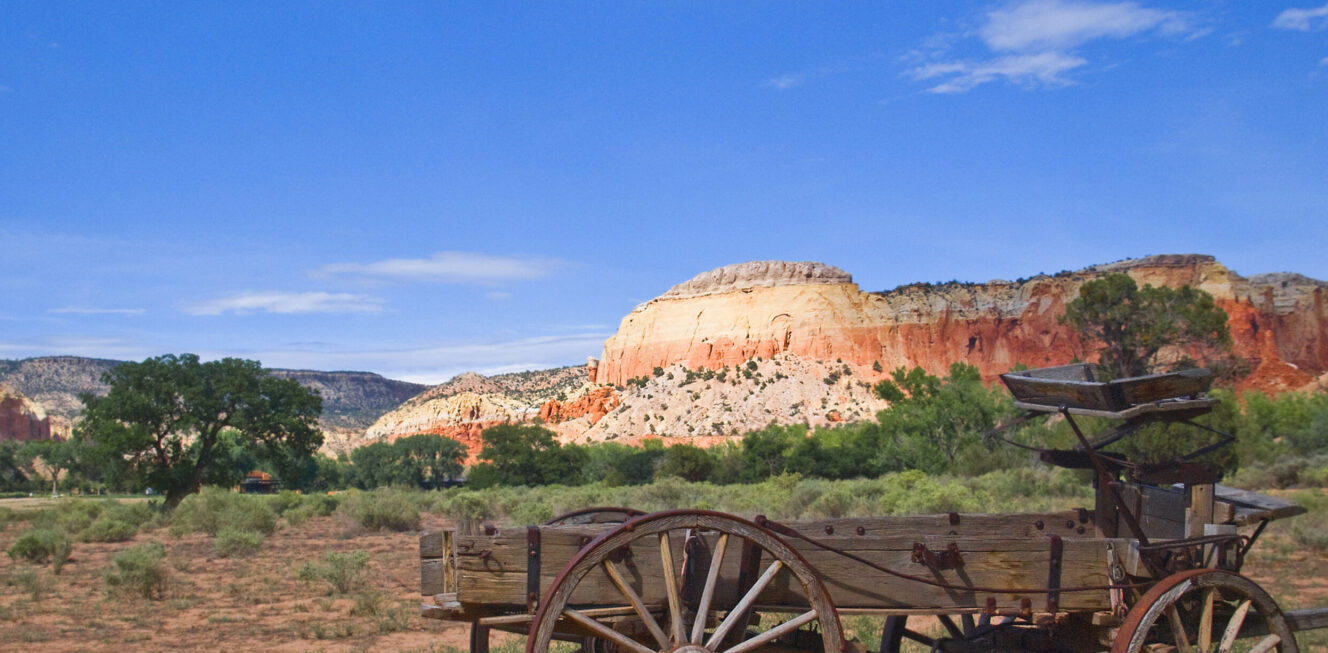 A rustic, weathered wooden wagon at the Buckboard area of Ghost Ranch in New Mexico, set against the scenic backdrop of red rock formations and piñon-juniper woodlands.