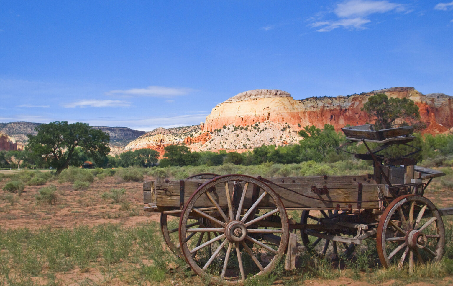 A rustic, weathered wooden wagon at the Buckboard area of Ghost Ranch in New Mexico, set against the scenic backdrop of red rock formations and piñon-juniper woodlands.