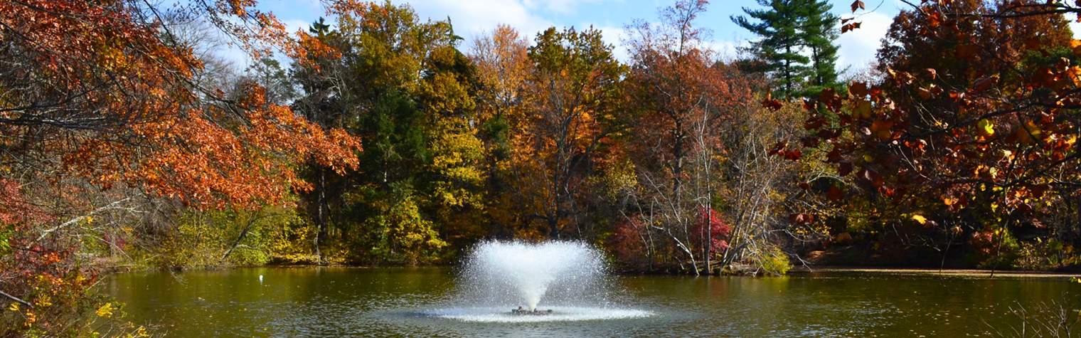 A serene water fountain sprays water into a tranquil lake, surrounded by vibrant autumn woodlands, creating a picturesque scene that combines the soothing sounds of water with the warm, golden hues of fall foliage.