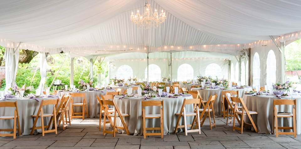 Elegant event setting at River Farm, featuring white-draped canopies adorned with beautiful chandeliers, surrounded by lush gardens. Tables are exquisitely set with lovely rose and lavender flower centerpieces, creating a serene and sophisticated atmosphere perfect for weddings and special occasions.