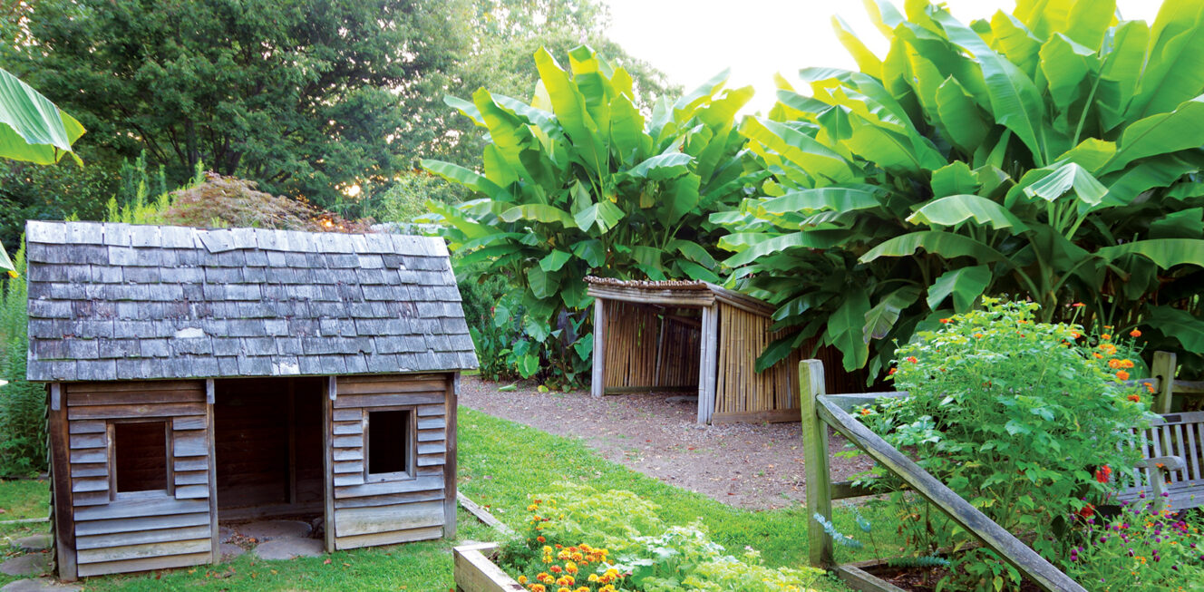 A child-sized wooden house nestled among tropical trees, lush green grass, and vibrant flowers at the Children's Garden at River Farm, providing a whimsical setting that sparks imagination and creativity in young minds.