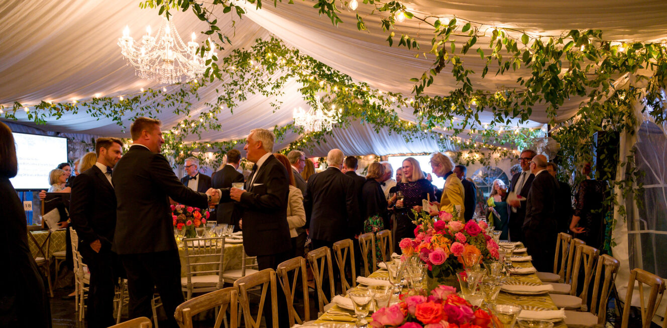 Guests mingle in suits and formal attire at an elegant event under a beautifully decorated tent adorned with ivy and a stunning hanging chandelier at River Farm. The setting features exquisite table arrangements with rose centerpieces, creating a sophisticated and romantic ambiance amidst the picturesque surroundings.