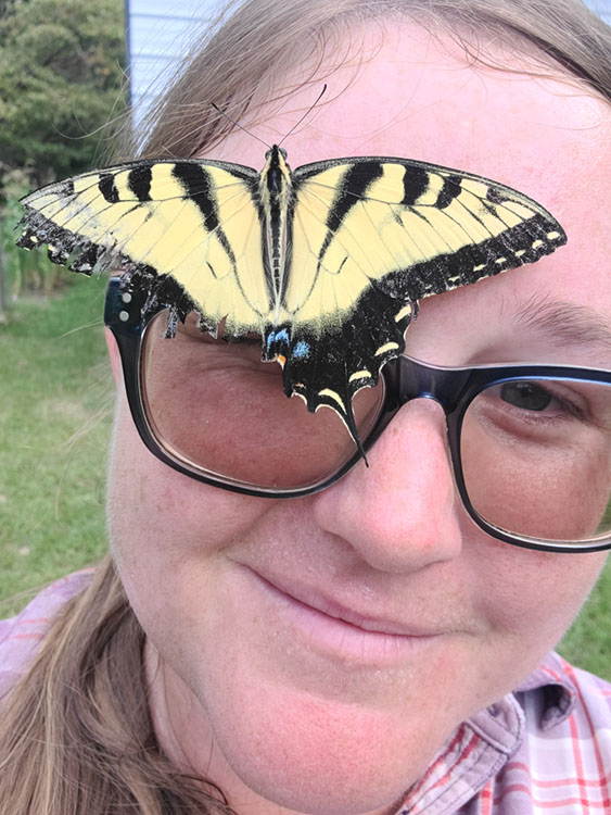 A big yellow butterfly has settled on the glasses frame of Amanda Bratcher, winner of the 2025 Emerging Horticultural Professional Award, part of the American Horticultural Society's Great American Gardeners Awards.