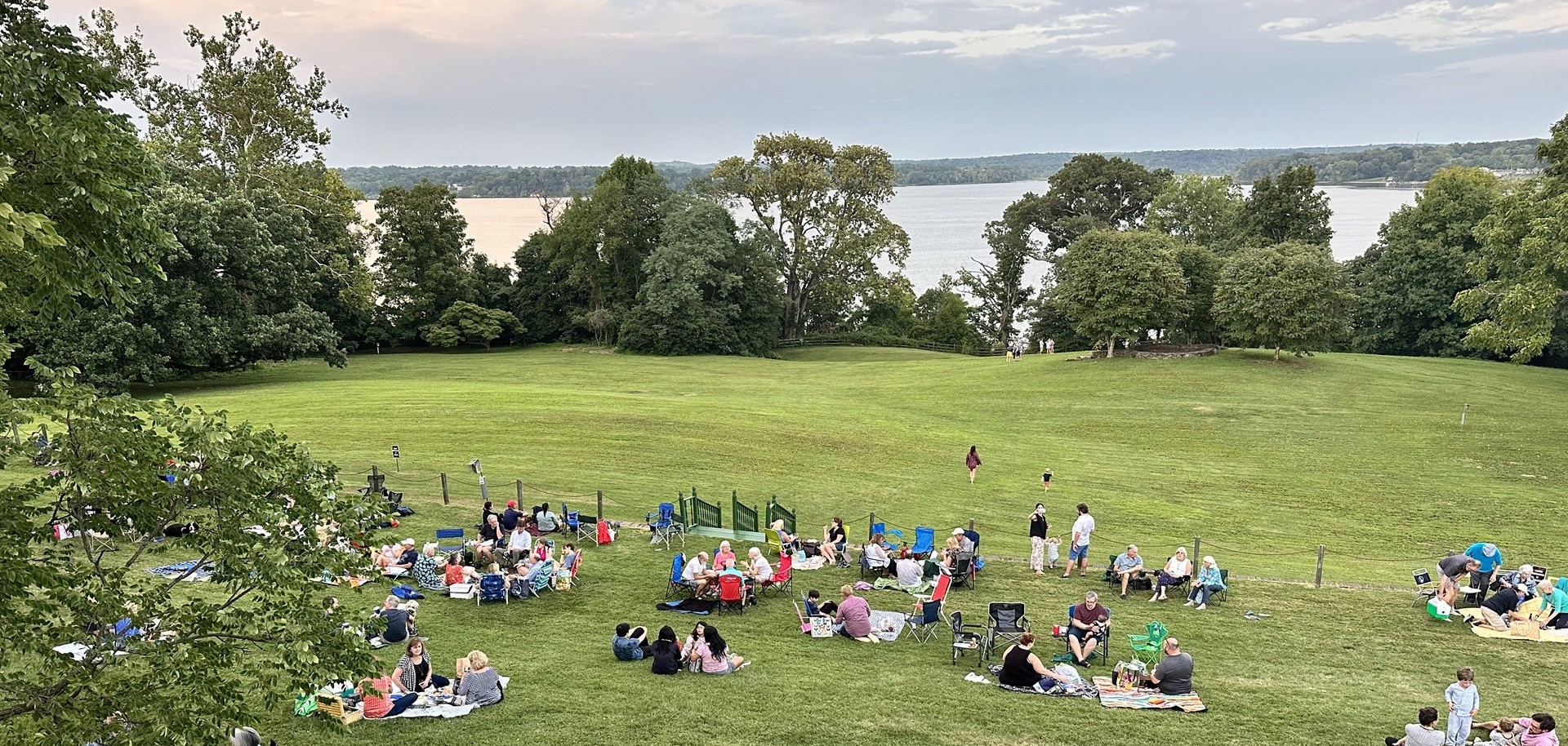 A crowd of people sit on a green lawn sloping down to a river. Large trees line the river bank under a sunset sky.