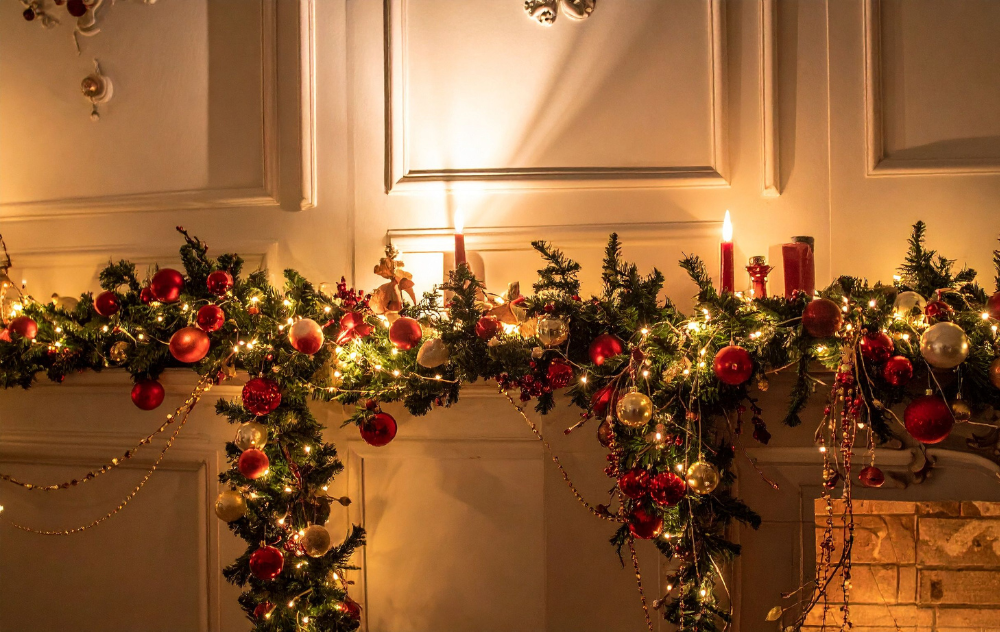 A green evergreen garland with red berries and white lights sits along a mantle against a cream colored wall with a fireplace in the bottom right corner.